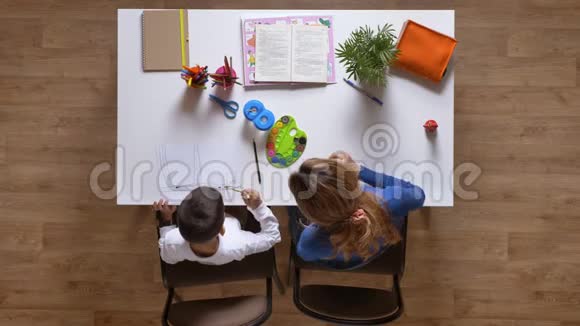 年轻的母亲和儿子一起画画坐在桌子后面孩子在做作业顶着镶木地板视频的预览图