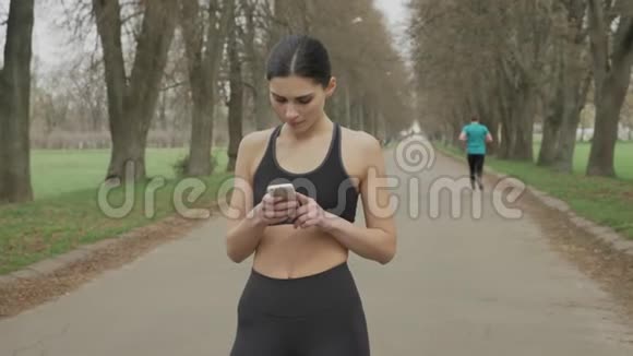年轻健康的女孩看着她的智能手机夏天在公园里跑步交流观念运动观念视频的预览图