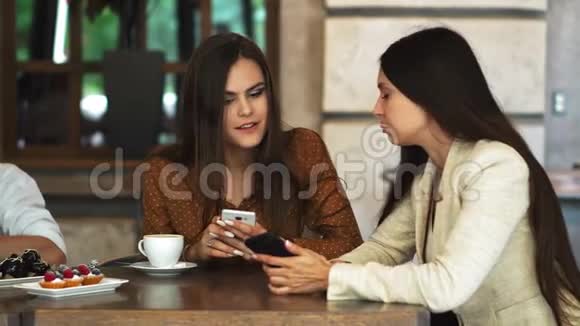 一对一会议两个年轻的女商人坐在咖啡馆的桌子旁女孩在智能手机屏幕上显示她的朋友形象视频的预览图