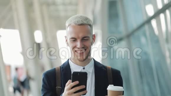 年轻有魅力的商人站在商务中心附近带着智能手机咖啡和耳机他看着视频的预览图