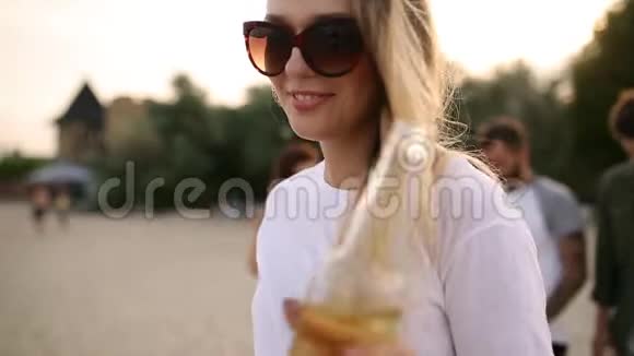 日落时分年轻漂亮的女人在海滩上散步喝着玻璃瓶慢镜头视频的预览图
