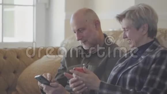 一对成年夫妇坐在大沙发上秃头男子在手机上为他的成熟女人展示照片她个子矮小视频的预览图