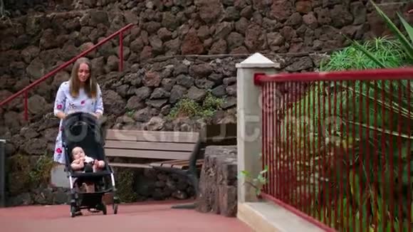 一位年轻的母亲带着一个婴儿坐在轮椅上行走在公园里那里有棕榈树和瀑布一个幸福的家庭视频的预览图