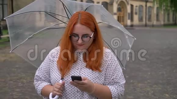 雨天公园里戴眼镜的胖姜姑娘在智能手机上打字笑着撑着伞视频的预览图