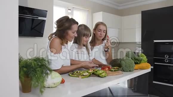 快乐的母亲和她的两个女儿正在家厨房做饭吃蔬菜食物健康饮食家庭视频的预览图