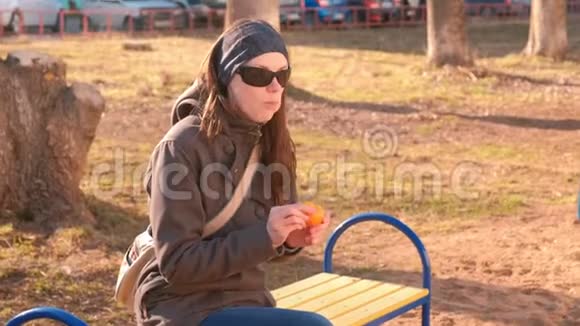 年轻的黑发女人戴着太阳镜在公园里坐在长凳上剥皮吃橘子视频的预览图