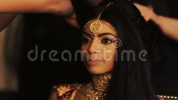 迷人的印度新娘把她娇嫩的手指放在太阳穴后面而女人则准备好她的头发视频的预览图