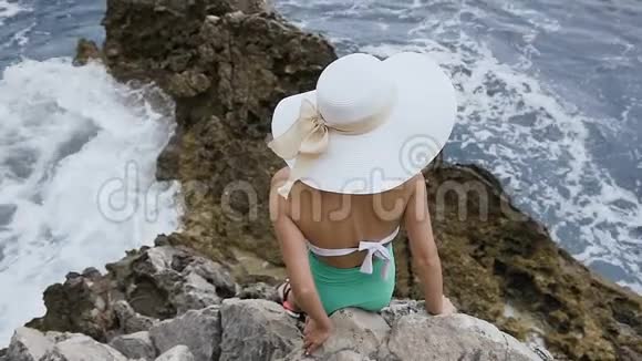 上景一个戴着帽子的年轻女孩站在岩石附近大海在那里波涛汹涌年轻的黑发女人戴着大帽子视频的预览图