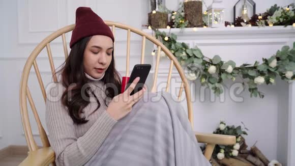 亚洲女性在客厅里躺在椅子上时使用智能手机进行短信阅读信息传递和在线购买视频的预览图