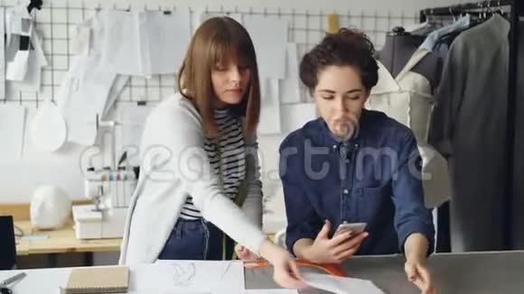 年轻的服装设计企业家正在看草图和检查智能手机有魅力的女人正在讨论视频的预览图