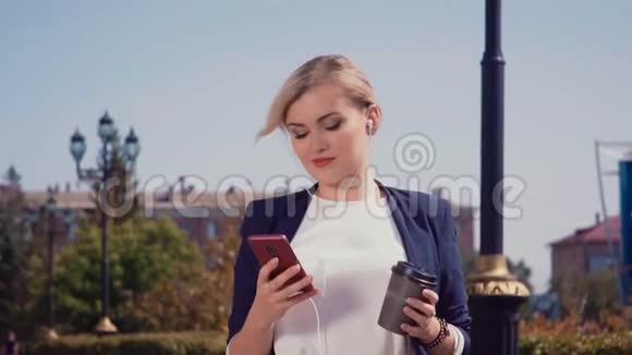 一个女孩站在阳光明媚的街道上拿着一部智能手机和一杯咖啡女孩正看着视频的预览图