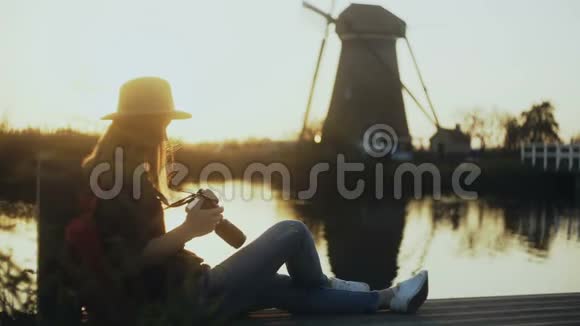 女摄影师坐在日落湖码头上带相机的女孩拍了一张传统乡村风车的照片4K视频的预览图