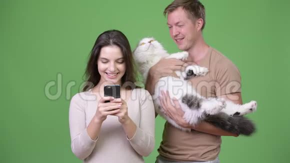 年轻夫妇在绿背景下和猫一起使用电话视频的预览图