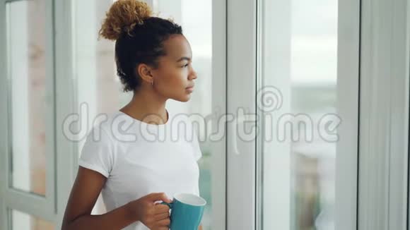 微笑的年轻女子正站在窗边喝茶看着外面享受着家里的闲暇时光视频的预览图