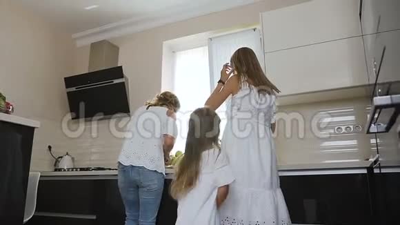 快乐的小女孩穿着白色连衣裙和姐姐和妈妈一起跳舞一边听音乐一边在厨房做饭视频的预览图