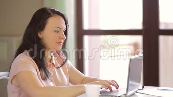 一个年轻的女人坐在咖啡馆里在笔记本电脑上工作喝着一次性杯子里的咖啡慢节奏视频的预览图