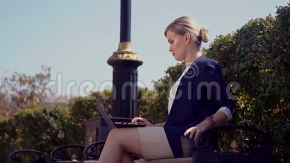 一个年轻的女孩坐在办公楼旁边的街道上在笔记本电脑后面工作笔记本电脑在她腿上视频的预览图