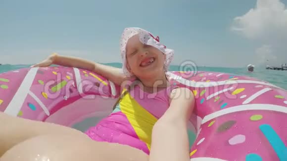 一个小女孩戴着一顶凉爽的帽子穿着一件鲜艳的比基尼骑在一个圆圈上自拍对着镜头微笑视频的预览图