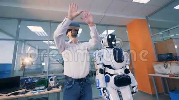 男工程师站在一个机器人旁边通过眼镜操纵虚拟现实视频的预览图