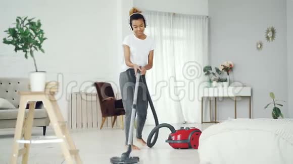 开朗的家庭主妇在家里打扫卫生时使用吸尘器通过耳机听音乐跳舞和视频的预览图