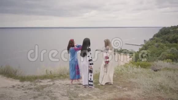 三个可爱的女朋友穿着夏天的长时装在田野上背景是湖或河二视频的预览图