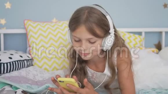 快乐的微笑女孩躺在床上带着智能手机和耳机在家听音乐视频的预览图