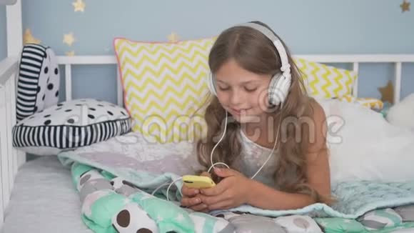 快乐的微笑女孩躺在床上带着智能手机和耳机在家听音乐视频的预览图
