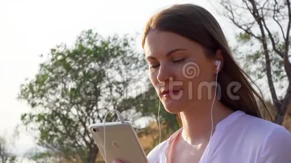 年轻快乐的女人在绿色公园里用手机女性通过手机慢动作听音乐视频的预览图