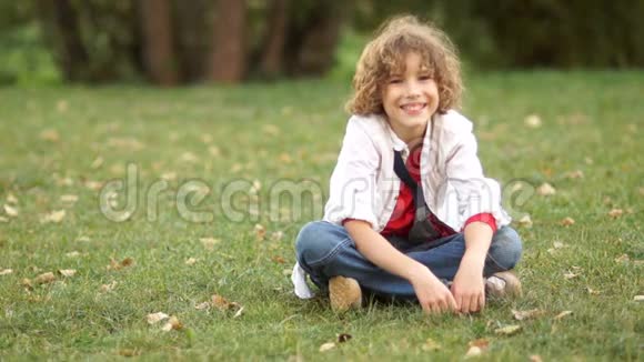 一个微笑的男孩坐在夏天公园的绿草上一个在度假的小学生穿着一件白色衬衫和一件红色T恤视频的预览图