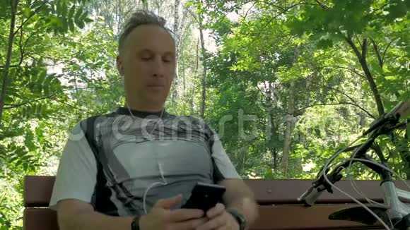 男运动员中年训练结束后在公园长椅上休息听智能手机上的音乐视频的预览图
