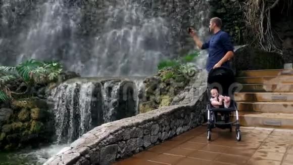 一位年轻的父亲和一个坐在轮椅上的孩子在瀑布附近散步他在智能手机上为社交拍照视频的预览图