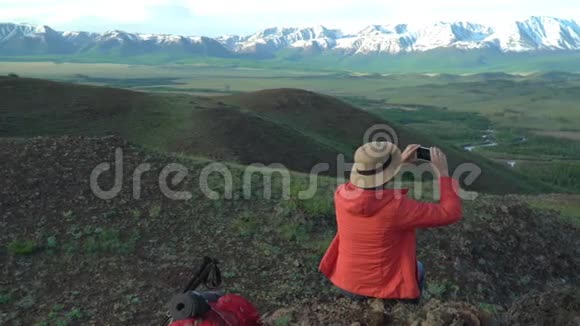 那个带着红色背包的旅行者的女人坐在那里拍摄雪山在女人身上穿着橙色夹克和视频的预览图