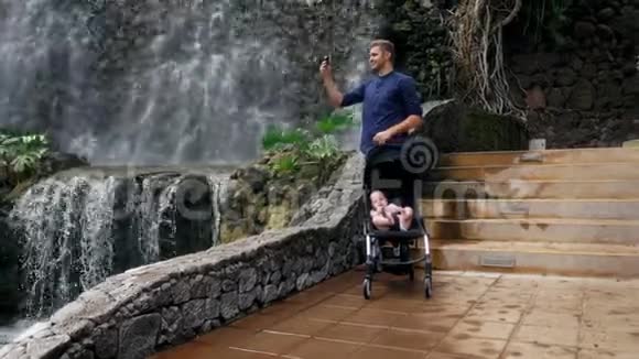 一位年轻的父亲和一个坐在轮椅上的孩子在瀑布附近散步他在智能手机上为社交拍照视频的预览图