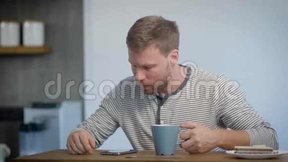 独自一人的年轻人正在他的公寓里吃早餐坐在厨房里喝着咖啡看着品视频的预览图