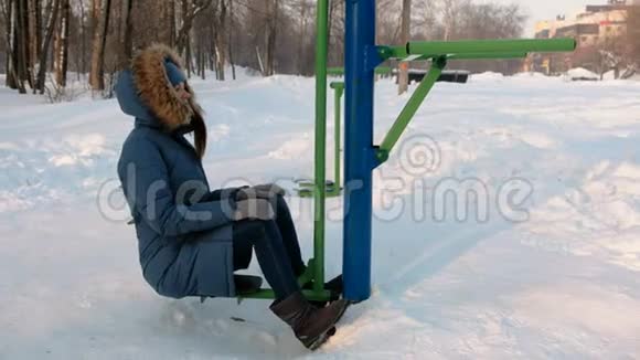 穿着带罩的蓝色羽绒服的女人正在冬季公园的模拟器上做腿部运动侧视视频的预览图