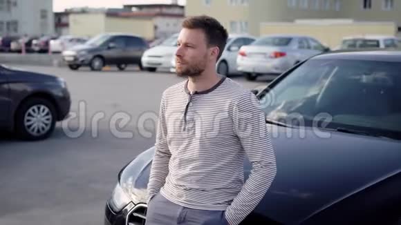 一个英俊的白种人坐在他的黑色大轿车的引擎盖上在夏季傍晚的停车区思考问题视频的预览图