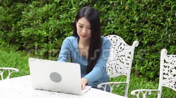 美丽的亚洲年轻女子微笑着坐在花园的椅子上在网上工作着笔记本电脑视频的预览图