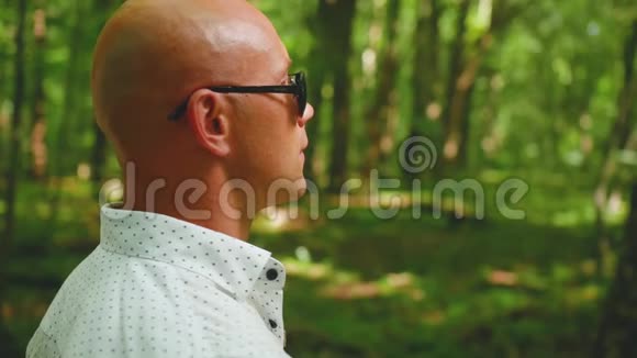 一个戴着太阳镜在森林里散步的秃头男人的肖像绿树夏天大自然近距离射击视频的预览图