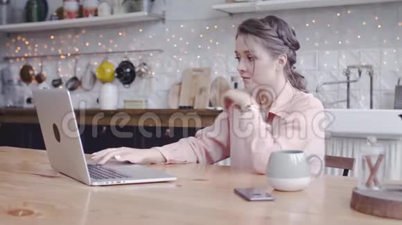 年轻漂亮的女人在家里厨房里用笔记本电脑工作库存录像漂亮的女人小心地在笔记本电脑上工作视频的预览图