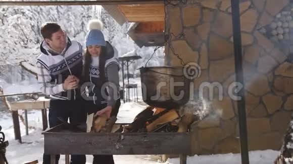 一对年轻快乐的夫妇在冬天的一天准备在一个木制森林房子的院子里明火视频的预览图
