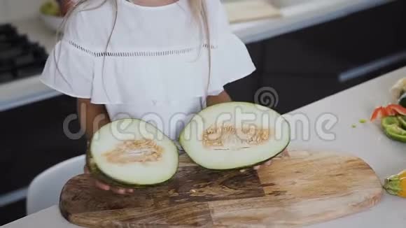 穿白衣服的女孩手里拿着一个瓜在家里的厨房里切成两半青瓜裂片的特写视频的预览图