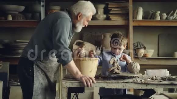 可爱的孩子正在他祖父的车间里玩粘土和陶工工具制作陶瓷玩具灰发胡子视频的预览图