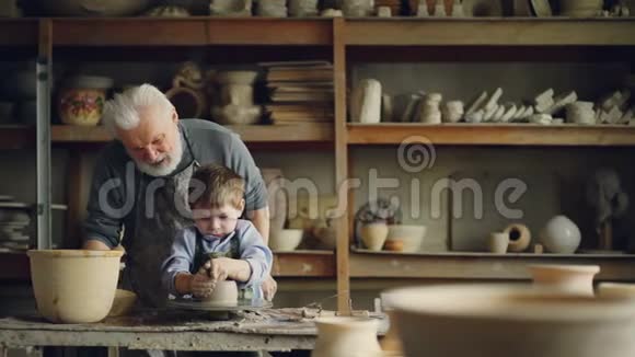 关心的祖父经验丰富的陶工正在教小男孩如何与粘土在陶工的车轮孙子在做视频的预览图