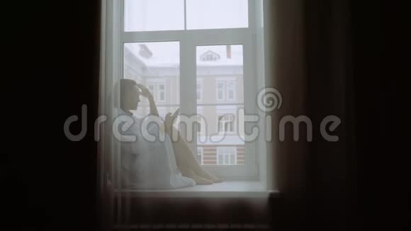 一个穿着白色外套手里拿着智能手机的女人坐在窗台上在冬天之外视频的预览图