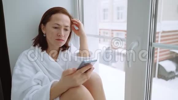 一个穿着白色外套手里拿着智能手机的女人坐在窗台上在冬天之外视频的预览图