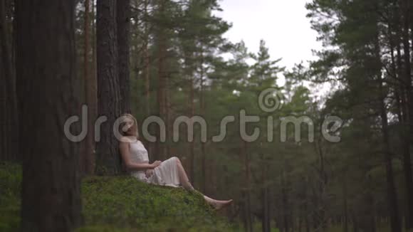 美丽的金发女郎坐在森林里的仙女穿着白色的裙子穿着常青的木头视频的预览图