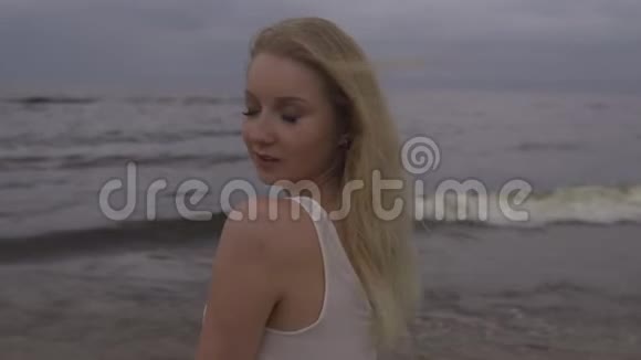 一幅美丽的金发美女海滩仙女的肖像穿着白色连衣裙在沉闷的阴郁中海浪拍打着海面视频的预览图