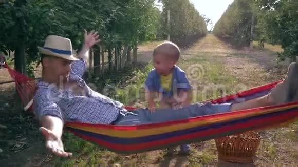快乐的孩子在苹果园水果篮附近的吊床上摇着他的父亲慢动作视频的预览图