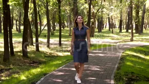 在公园里散步的年轻女人心情好的女孩微笑着在城市公园散步沿着这条路走来视频的预览图