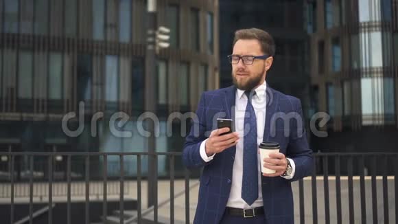 优雅的老板正在使用智能手机拿着咖啡站在办公楼的背景上视频的预览图